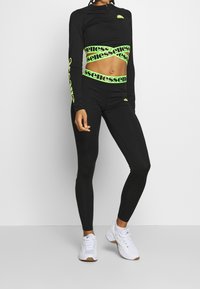 Svart långärmad topp med neongröna logotyper, kombinerad med högmidjade leggings med liknande neondetaljer. Vita sneakers fulländar stilen.