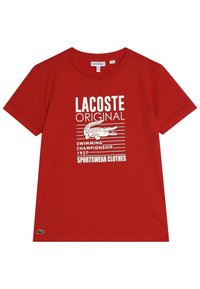 T-shirt rossa in cotone con maniche corte. Presenta testo bianco e un logo di coccodrillo, insieme a strisce orizzontali e un design a tema sportivo.