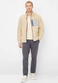 Fleecejacke in Beige mit einer grauen Brusttasche mit Reißverschluss, kombiniert mit grauer Hose und beigen Turnschuhen. Weiche Textur, legerer Schnitt.