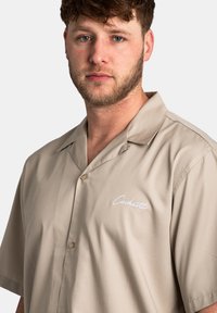Chemise beige à manches courtes en tissu lisse, dotée d'un design boutonné et d'un logo brodé blanc sur la poitrine.