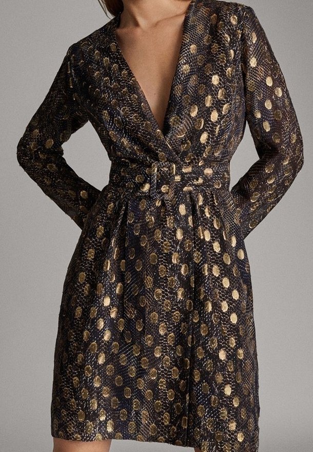 Femme portant une robe à manches longues, décolleté en V profond, motif à pois noirs et dorés, et une ceinture assortie à la taille.
