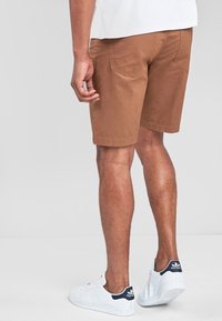 Shorts en coton marron à coupe droite, poches arrière et ourlets retournés, associés à des baskets blanches. Coupe ample et design décontracté.
