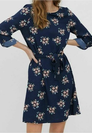 Frau trägt ein marineblaues Blumenkleid mit Dreiviertelärmeln und einem gebundenen Taillengürtel, steht mit den Händen in der Hüfte.