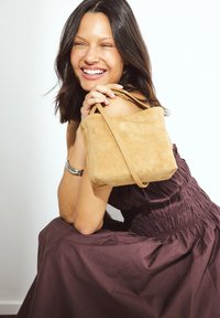 Borsa in suede di colore biscotto morbido, forma rettangolare, con manici corti e robusti e dettagli di cucitura minimi. Indossata con un abito scuro e arricciato.