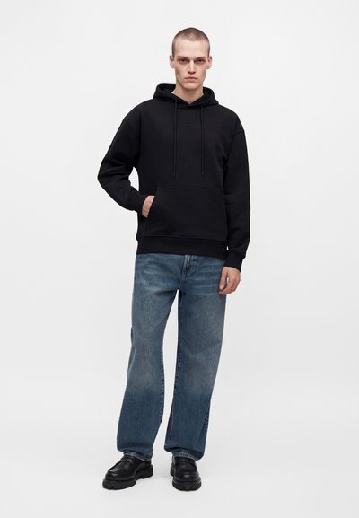 Jeune homme portant un sweat à capuche noir, un jean bleu large et des chaussures noires, debout devant un fond clair uni.