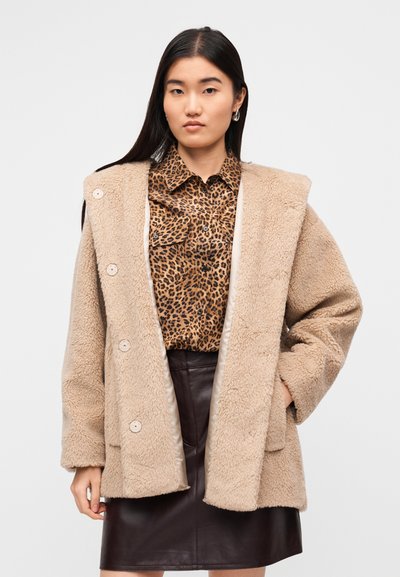 Μπεζ, υφασμάτινο, υπερμεγέθες παλτό με μεγάλα κουμπιά. Φοριέται πάνω από πουκάμισο με λεοπάρ σχέδιο και καφέ δερμάτινη φούστα.