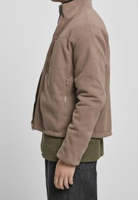 Giacca in pile marrone con colletto alto, chiusura con zip e tasche laterali. Indossata sopra un maglione a maglia verde con texture, abbinata a jeans scuri.