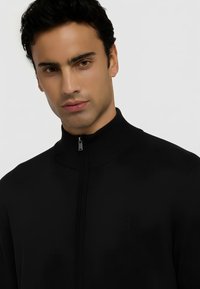 Černá zipová bunda s vysokým límcem, žebrovanou texturou a jemným logem na hrudi. Hladká tkanina s přiléhavým střihem.