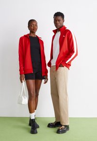 Røde sporty jakker med hvide sidelister, sort top, sorte shorts og beige bukser. Sorte sko, hvid taske. Glat tekstur, afslappet design.