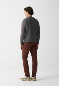 Hanorac gri cu manșete striate, purtat cu pantaloni maro. Adidașii au un design multicolor și o talpă texturată. Vedere din spate.