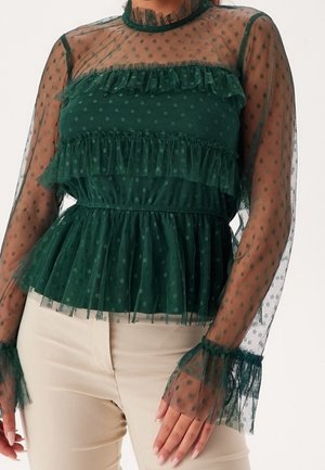 Blouse en maille transparente vert foncé à pois avec volants sur la poitrine, les poignets et la taille, associée à un pantalon beige.
