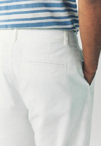 Valged linast püksid, millel on tagatasku, sile tekstuur ja lõdvestunud lõige. Kombineeritud sinise ja valge triibulise kootud topiga.