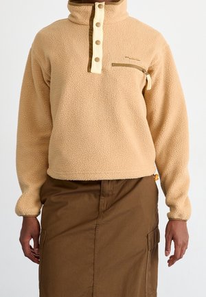 Pull polaire beige avec patte à boutons-pression et poche poitrine zippée porté avec une jupe cargo marron sur une personne debout.