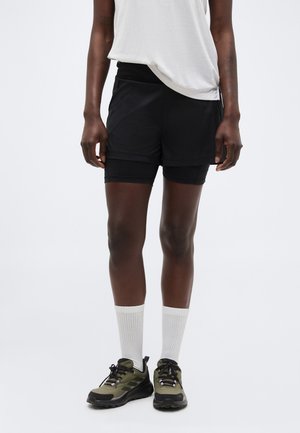 Persoană care poartă pantaloni scurți de alergare negri stratificați, tricou alb, șosete albe și pantofi sport verde-olive și negri.