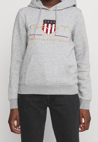 Felpa con cappuccio grigia in tessuto misto cotone, con tasca frontale, cappuccio con coulisse e logo ricamato con accenti rossi e dorati.