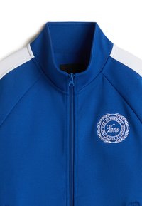Blaue Zip-Up-Trainerjacke mit hohem Kragen, weißen Schulterstreifen, strukturiertem Stoff und gesticktem Logo in kreisförmigem Design auf der Brust.