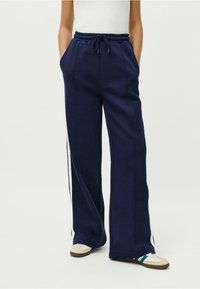 Jūras zila sporta bikses ar auklu jostasvietā, sānu kabatām un baltām svītrām gar kājām. Izgatavotas no mīksta, elastīga materiāla.