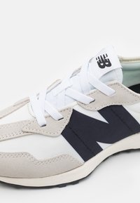 Lys grå og hvit sneaker med en marineblå logo, teksturerte semskede detaljer, og flate hvite lisser, med en gummisåle for bedre grep.