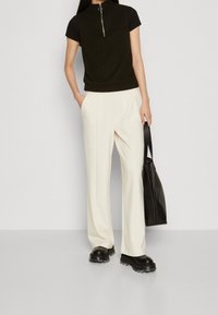 Ärmelloses Stricktop mit Reißverschluss, dunkelgrün; weite cremefarbene Hose; schwarze strukturierte Tasche; klobige schwarze Schuhe mit transparenten Akzenten.