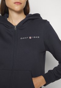 Femme portant un sweat à capuche zippé bleu marine avec le logo "GANT 1949" et un emblème de bouclier sur la poitrine, main dans la poche avant.