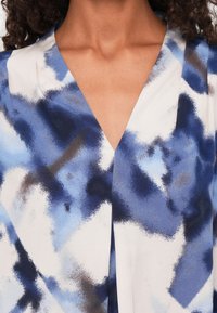 Blaue und weiße gemusterte Bluse mit tiefem V-Ausschnitt, ausgestattet mit abstrakten Aquarellsprühen und einer glatten Stofftextur.