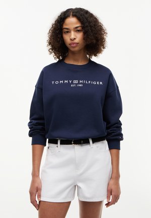 Jeune femme aux cheveux bouclés portant un sweat-shirt bleu marine Tommy Hilfiger et un short blanc sur un fond uni.