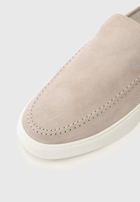 Suede slip-on sko i lys beige med hvit gummisåle. Har synlig søm langs den øvre kanten og rund tådesign.