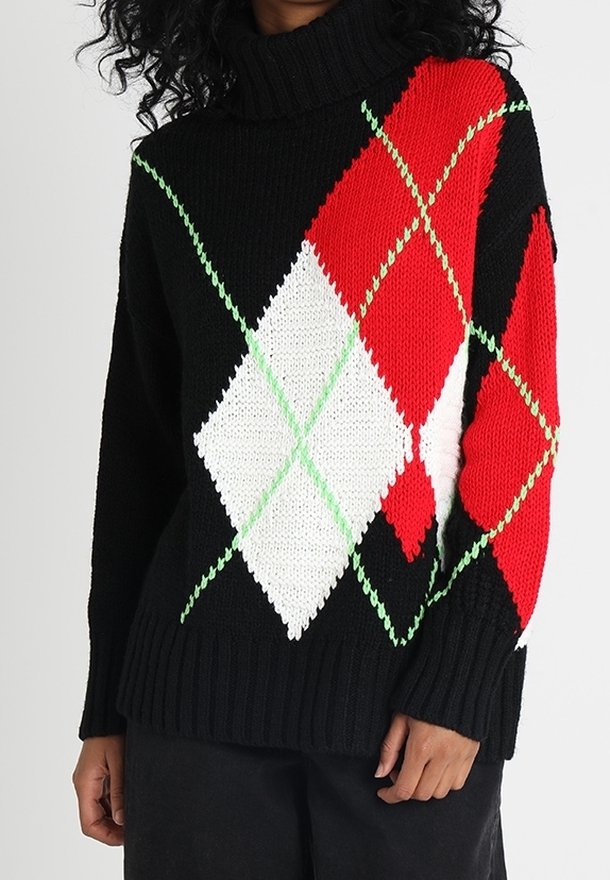 Pull à col roulé noir avec de grands motifs losange argyle rouges, blancs et verts porté par une personne aux cheveux bouclés foncés.