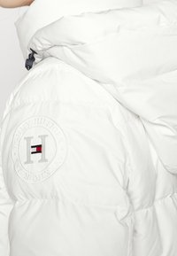 Vit puffjacka med en rund broderad logotyp med "Tommy Hilfiger" och ett centralt rödblått rand. Mjuk, quiltad textur.