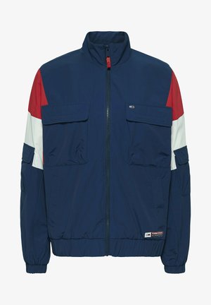 Jachetă de vânt de marină cu accente roșii și albe, două buzunare frontale, manșete și tiv elastic, și guler înalt. Material respirabil.