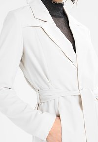 Biały, teksturowany długi płaszcz z kołnierzem i paskiem w talii, charakteryzujący się eleganckim designem i dopasowanymi rękawami.