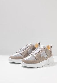 Szare zamszowe trampki z siateczkowymi panelami, białymi paskami i grubymi białymi podeszwami. Designerski wygląd dopełniają sznurowadła oraz pętla z tyłu buta.