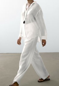 Camicia bianca in lino leggero con maniche lunghe e pantaloni abbinati dal taglio rilassato. Collana con pendente nera, sandali piatti; texture liscia.