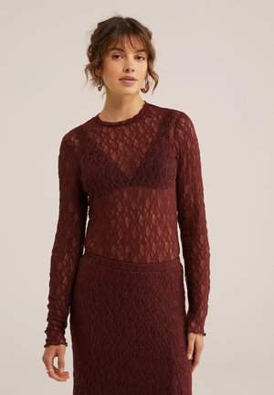 Žena so svetlohnedými vlasmi upravenými do drdolu, oblečená v dlhom rukáve bordového čipkovaného topu a zodpovedajúcej sukni na jednoduchom pozadí.