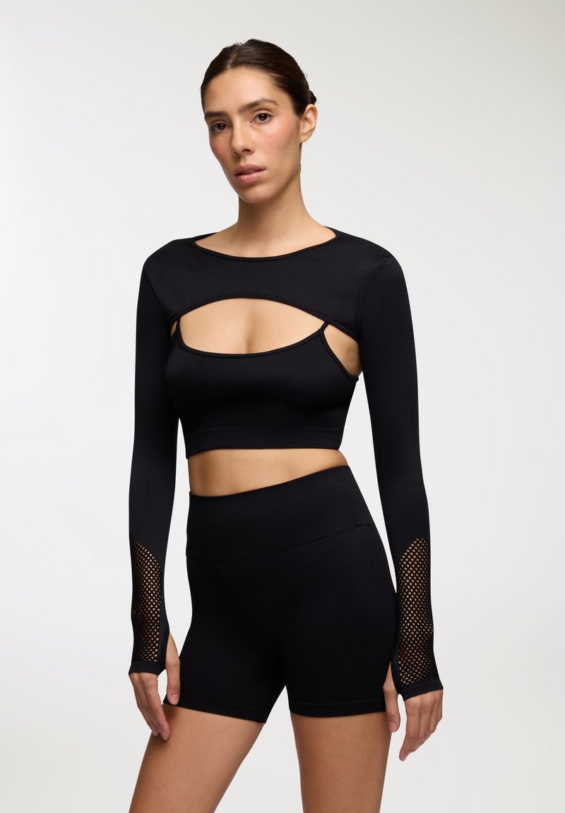 Czarny zestaw sportowy zawierający bluzkę z długim rękawem z wycięciami i siateczkowymi akcentami na rękawach, w połączeniu z szortami o wysokim stanie. Gładka tekstura.