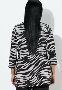 Kobieta z długimi, prostymi czarnymi włosami, ubrana w czarną i białą bluzkę w paski zebry, stojąca na tle białego, jednolitego tła.