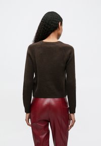Donna con lunghi capelli ricci, che indossa un maglione di maglia marrone scuro e pantaloni di pelle rossa a vita alta, vista di schiena su uno sfondo neutro.