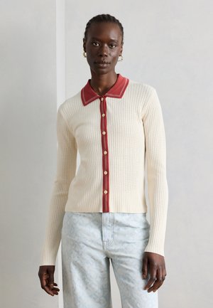 Femme portant un cardigan en maille crème avec un col rouge et une patte de bouton, associé à un pantalon bleu clair à motifs, debout contre un mur uni.