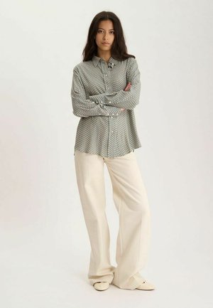 Jeune femme debout, les bras croisés, portant une chemise à manches longues à motifs et un pantalon large crème sur fond uni.