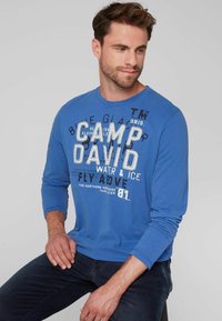 Niebieska longsleeve z napisem "CAMP DAVID" w białym i czarnym kolorze. Miękki materiał, okrągły dekolt i luźny krój.