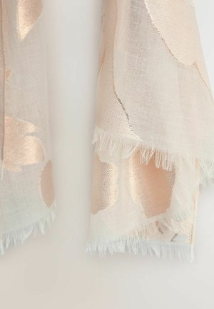 Lysebeige stof med store, bløde gyldne blomsterprint og frynsekant, der hænger mod en ensartet hvid baggrund.