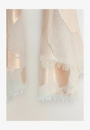 Világos bézs szövet nagy, puha arany virágmintákkal és rojtos szegéllyel, fehér, sima háttér előtt függve.