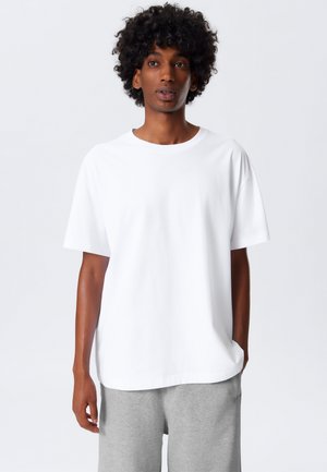 Weißes T-Shirt mit kurzen Ärmeln aus weichem Stoff, mit einem runden Halsausschnitt und lockerer Passform, kombiniert mit grauen Shorts.