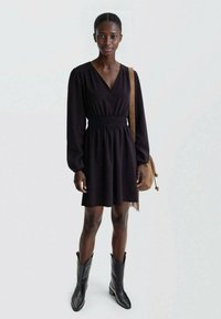 Črna kratka obleka z globokim V-izrezom, nabrano pasom, dolgimi rokavi in razširjenim krilom, kombinirana s črnimi kavbojskimi čevlji in rjavim nahrbtnikom.