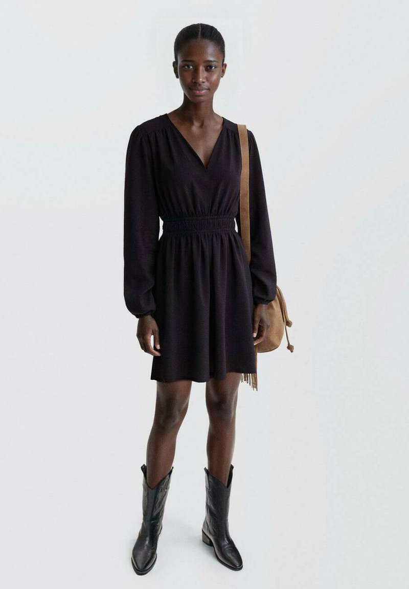 Črna kratka obleka z globokim V-izrezom, nabrano pasom, dolgimi rokavi in razširjenim krilom, kombinirana s črnimi kavbojskimi čevlji in rjavim nahrbtnikom.