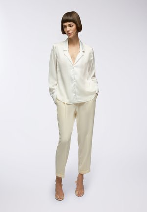 Blusa in satin avorio con una leggera lucentezza, maniche lunghe, chiusura con bottoni e colletto a punta abbinata a pantaloni crema su misura.
