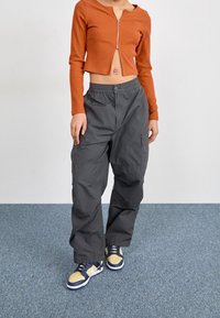Pantalones cargo grises con bolsillos, combinados con un top corto acanalado naranja con cremallera, y zapatillas amarillas/azules sobre un suelo azul texturizado.
