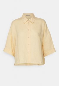 Lyse beige løs pasform linnedshirt med korte brede ærmer, knaplukning foran og klassisk krave mod en ensfarvet baggrund.