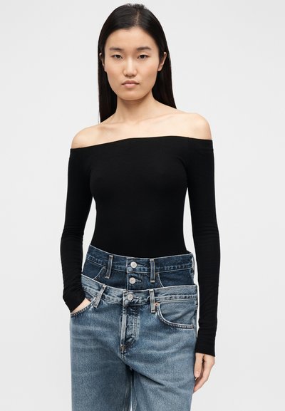 Μαύρο τοπ με μακριά μανίκια και ακάλυπτους ώμους, συνδυασμένο με ψηλόμεσο μπλε τζιν με πολλαπλά κουμπιά και τσέπες, με λείο ύφασμα.