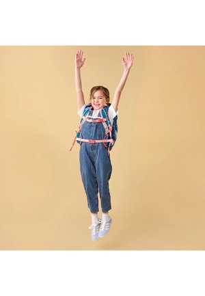 Young girl in striped overalls and sneakers jumps with arms raised, wearing a blue and pink backpack against beige background.